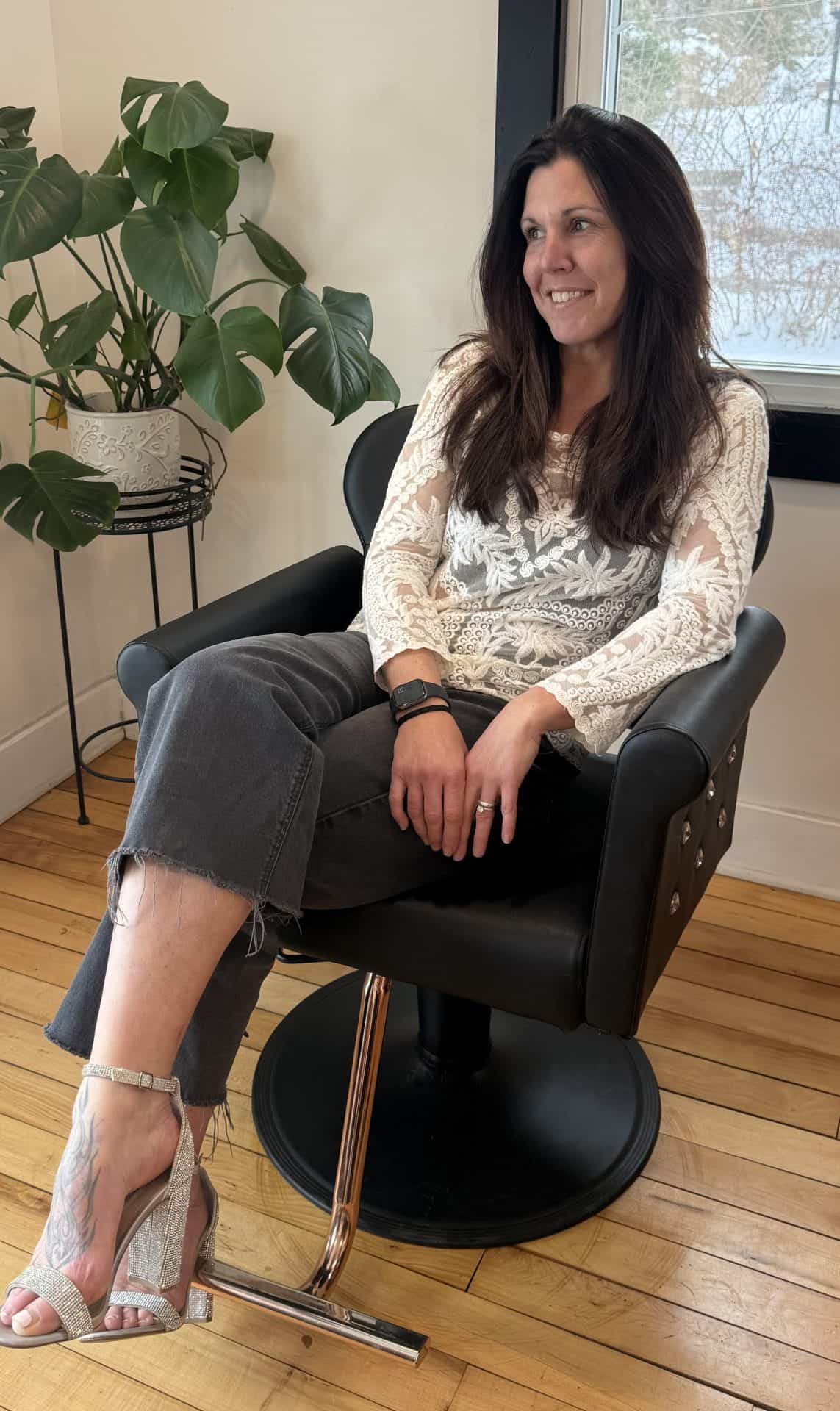 A woman with long hair is sitting comfortably in a stylish black chair, wearing a light sweater and relaxed pants, smiling in a well-lit room.